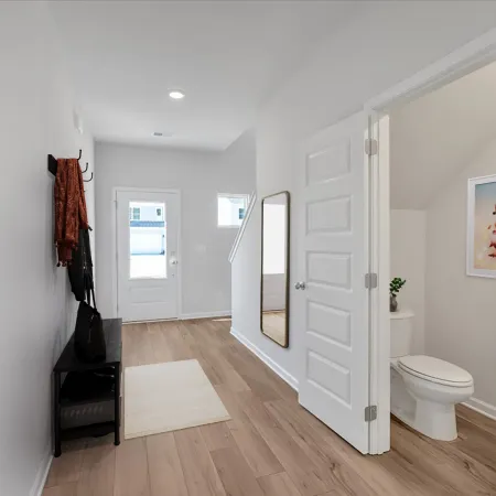 Bright hallway with wooden floor, a mirror on the wall, a small bench, and an open door to a bathroom.