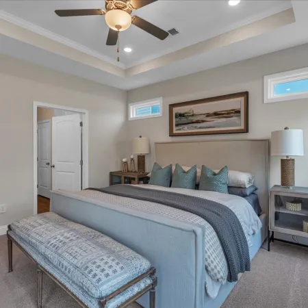 Modern bedroom with a king-size bed, blue and gray bedding, bench, lamps, and ceiling fan under tray ceiling.