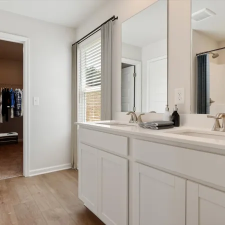 Modern bathroom with white double sink vanity, large mirror, wood flooring, and adjacent walk-in closet with hanging clothes.