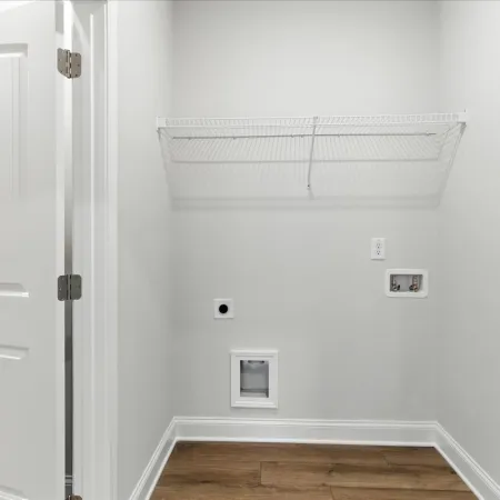Empty laundry room with white walls, wood floor, wire shelf, and hookups for washer and dryer installation.