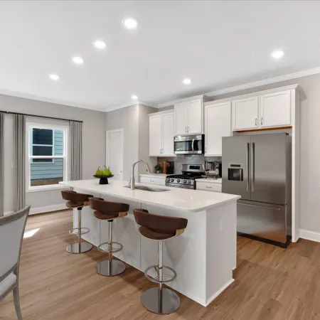Modern white kitchen with island, stainless steel appliances, wooden bar stools, and dining area with natural light.