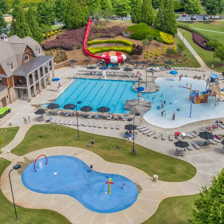 Aerial view of a public pool complex with swimming pool, water play area, slide, and splash pad, surrounded by green parkland.