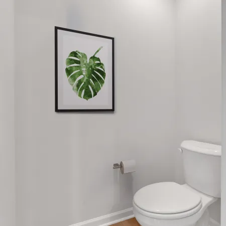 Minimalist bathroom corner with white toilet, wooden floor, leaf wall art, and toilet paper holder.