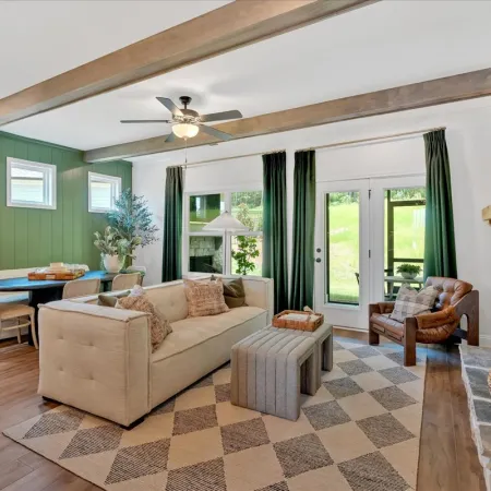 Bright living room with beige sofa, patterned rug, green accent wall, stone fireplace, and large windows with green curtains
