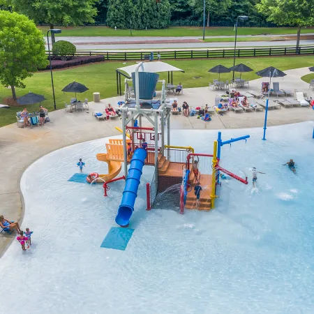Outdoor water park with a splash pad, slides, and people enjoying in shallow pool under umbrellas.