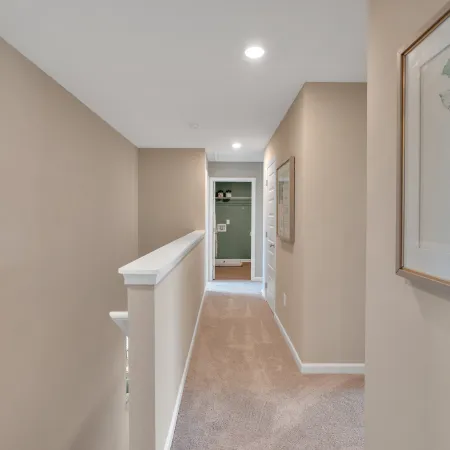 Bright hallway with neutral walls, carpet flooring, and framed artwork, leading to a green-themed room.