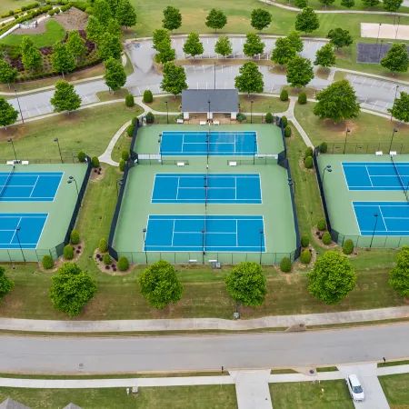 Aerial view of six blue and green tennis courts in a park surrounded by trees and a nearby playground area.