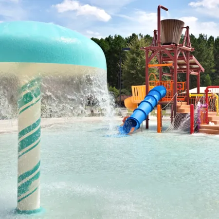 Water park splash pad with large mushroom fountain and colorful play structures under blue sky.