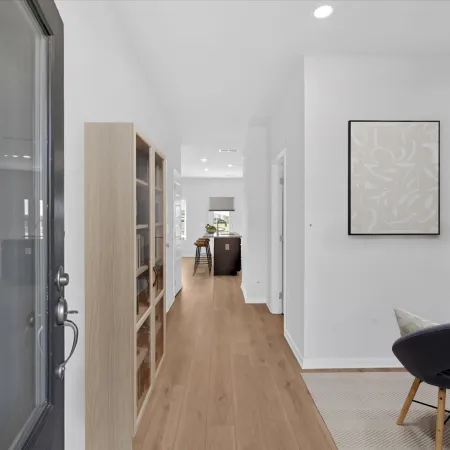 Bright modern hallway with wooden flooring, glass cabinet, desk area, and open kitchen in the background