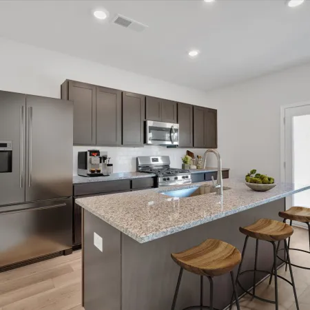 Modern kitchen with granite island, stainless steel appliances, dark cabinets, and three wooden bar stools under bright lighting.
