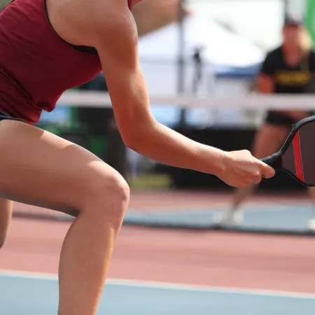 Athlete in red top and gray shorts playing pickleball hitting yellow ball with paddle on court.