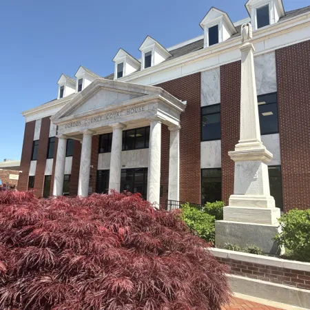 Gordon County Courthouse with brick facade, white columns, red foliage, and a stone monument under blue sky.