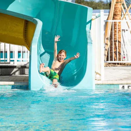 Happy boy in green shorts sliding down a blue water slide into a pool on a sunny day.