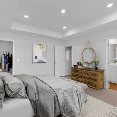 Modern bedroom with gray bedding, wooden dresser, round mirror, and connected bathroom with white vanity.