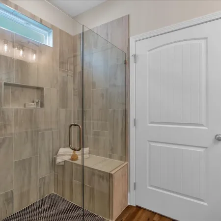 Modern bathroom with glass shower enclosure, built-in bench, wood-look tiles, and white door.