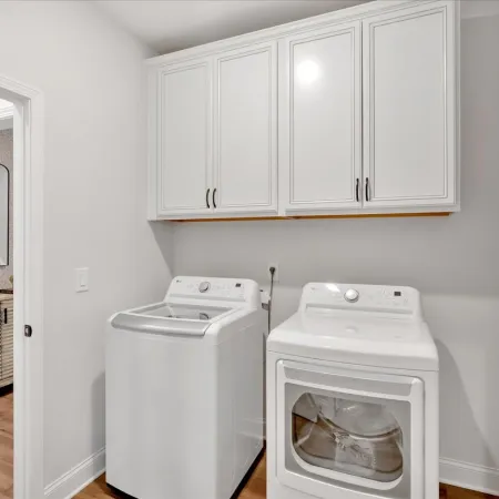 Bright laundry room with white washer and dryer under white cabinets and a glimpse into adjacent living space.