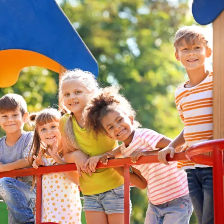 Group of five happy children leaning on playground equipment under a sunny sky with greenery in the background