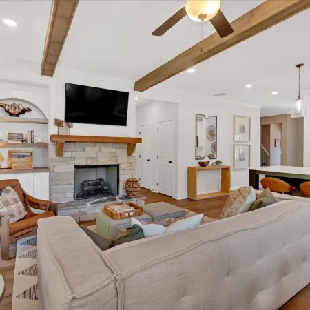 Bright living room with beige sofa, stone fireplace, wall-mounted TV, and adjacent kitchen with bar stools.