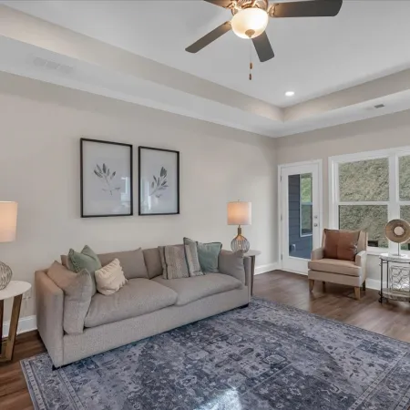 Modern living room with beige sofa, two armchairs, lamps, ceiling fan, and large window with natural light