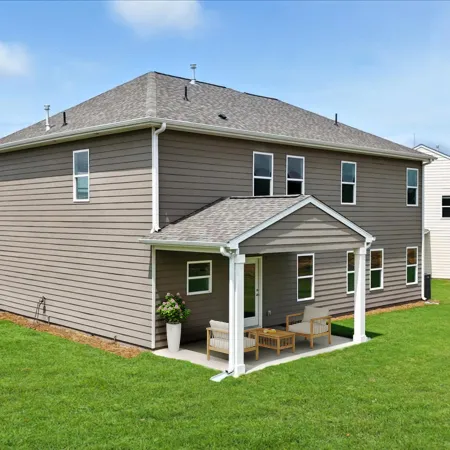 Two-story beige suburban house with covered patio and outdoor furniture on a green lawn under blue sky.