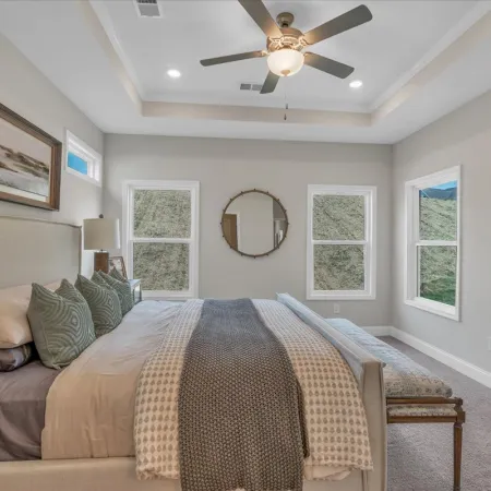 Modern bedroom with beige walls, large bed, patterned blankets, green pillows, ceiling fan, and three windows.