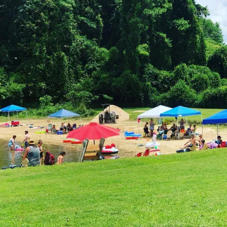 People enjoy a sunny day at a sandy beach with umbrellas, tents, and green trees in the background