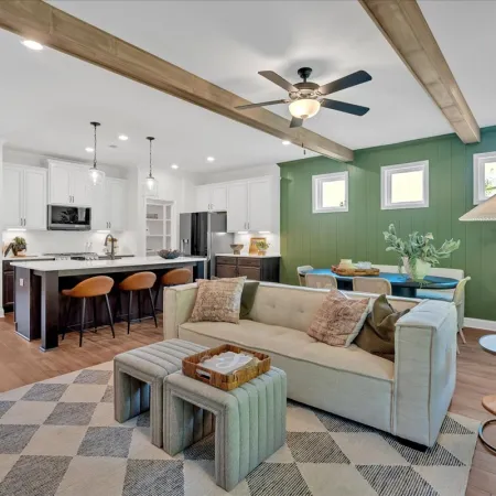 Open concept living room and kitchen with green accent wall, beige sofa, wooden beams, and modern lighting fixtures.