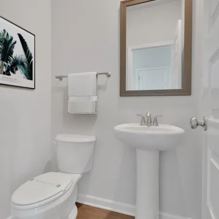 Bright small bathroom with white toilet, pedestal sink, large mirror, towel rack, and wall art on wood floor.