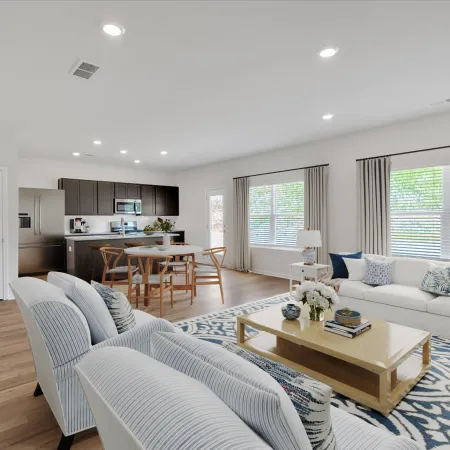 Bright open-concept living room and kitchen with white sofas, wooden dining table, and large windows with curtains.