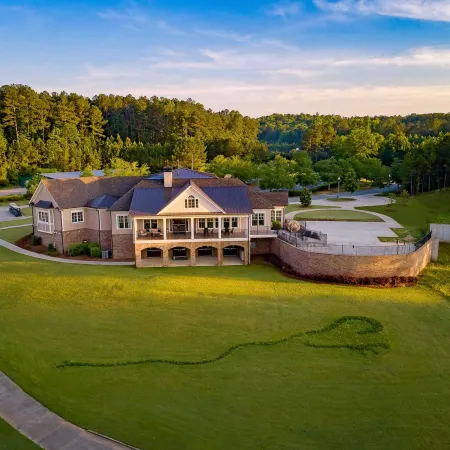 A large house overlooks a golf course with a flag-shaped grass pattern surrounded by trees under a clear sky.