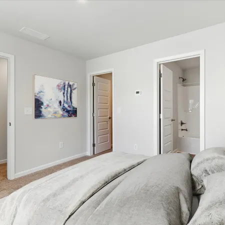 Modern bedroom with gray bedding, white walls, open doors leading to a hallway and bathroom with a bathtub.