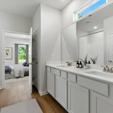Bright modern bathroom with double vanity, large mirror, white cabinets, wood flooring, and view into bedroom.