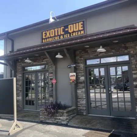 Exterior of Exotic-Que Barbecue & Ice Cream restaurant with stone facade and glass double doors under bright daylight.