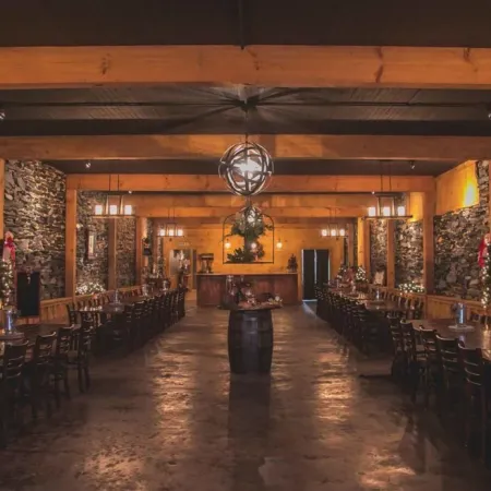 Rustic dining hall with stone walls, wooden beams, long tables, chairs, and warm ambient lighting.