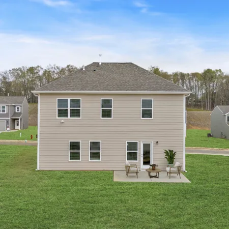 Back view of beige two-story house with patio seating on green lawn in suburban neighborhood under blue sky