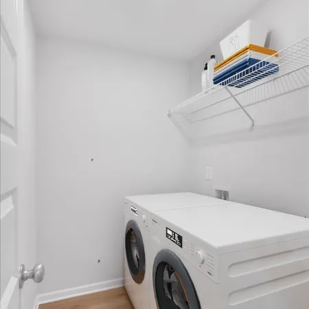 Bright laundry room with front-load washer and dryer, white walls, wire shelf with folded towels and detergent bottles.