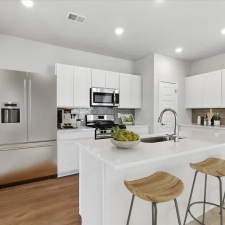 Modern white kitchen with stainless steel appliances, large island, wooden bar stools, and hardwood floor.