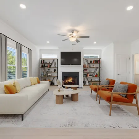 Bright modern living room with large windows, white sofa, orange chairs, fireplace, and wooden coffee tables.