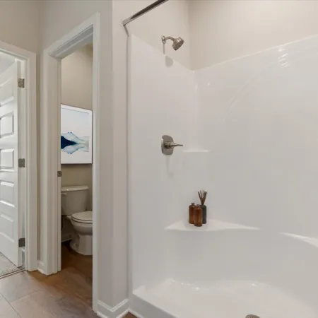 Bright bathroom with white bathtub, shower, and view into carpeted bedroom and toilet room with artwork.