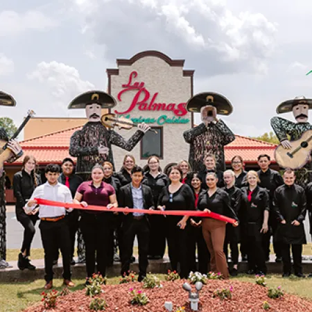 Group of people holding a red ribbon for grand opening in front of La Palmas Mexican Cuisine with mariachi statues.