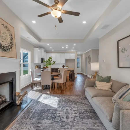 Bright open living room with fireplace, ceiling fan, gray sofa, dining table, and hardwood flooring under natural light