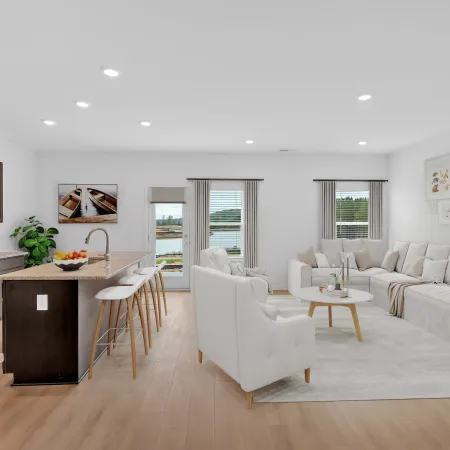 Modern open-concept living room and kitchen with white furniture, wood flooring, and large windows overlooking a lake.