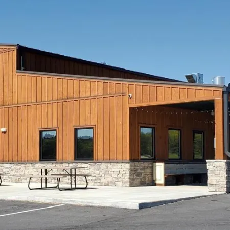Modern rustic wooden building with Pizza Farm sign, outdoor seating, and American flag on a clear day.