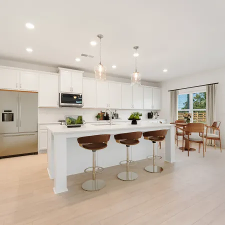 Bright modern kitchen with white cabinetry, large island with three brown stools, stainless steel appliances, and dining area.
