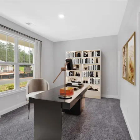 Modern home office with large window, gray carpet, black desk, beige chair, white bookshelf, and framed wall art