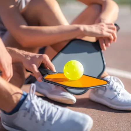 Two people in sportswear preparing to play pickleball with a yellow ball on a court surface.