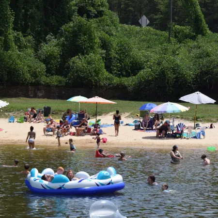 Crowded sandy beach with colorful umbrellas, people swimming, and children playing in lake water on a sunny day.