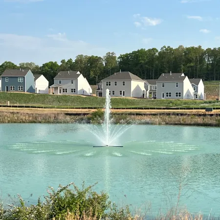 Tranquil pond with fountain surrounded by modern homes and greenery under a clear blue sky.