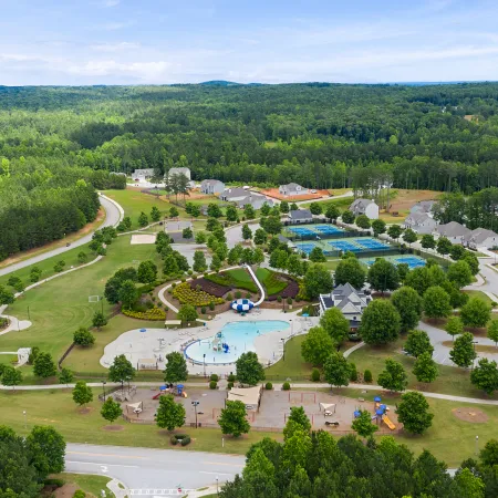 Aerial view of a suburban community park with pools, tennis courts, playgrounds, houses, and surrounding woods under a blue sky.