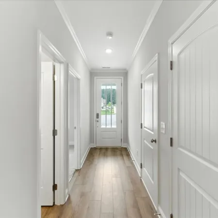 Bright hallway with white walls, wooden flooring, multiple white doors, and a glass-paneled front door.